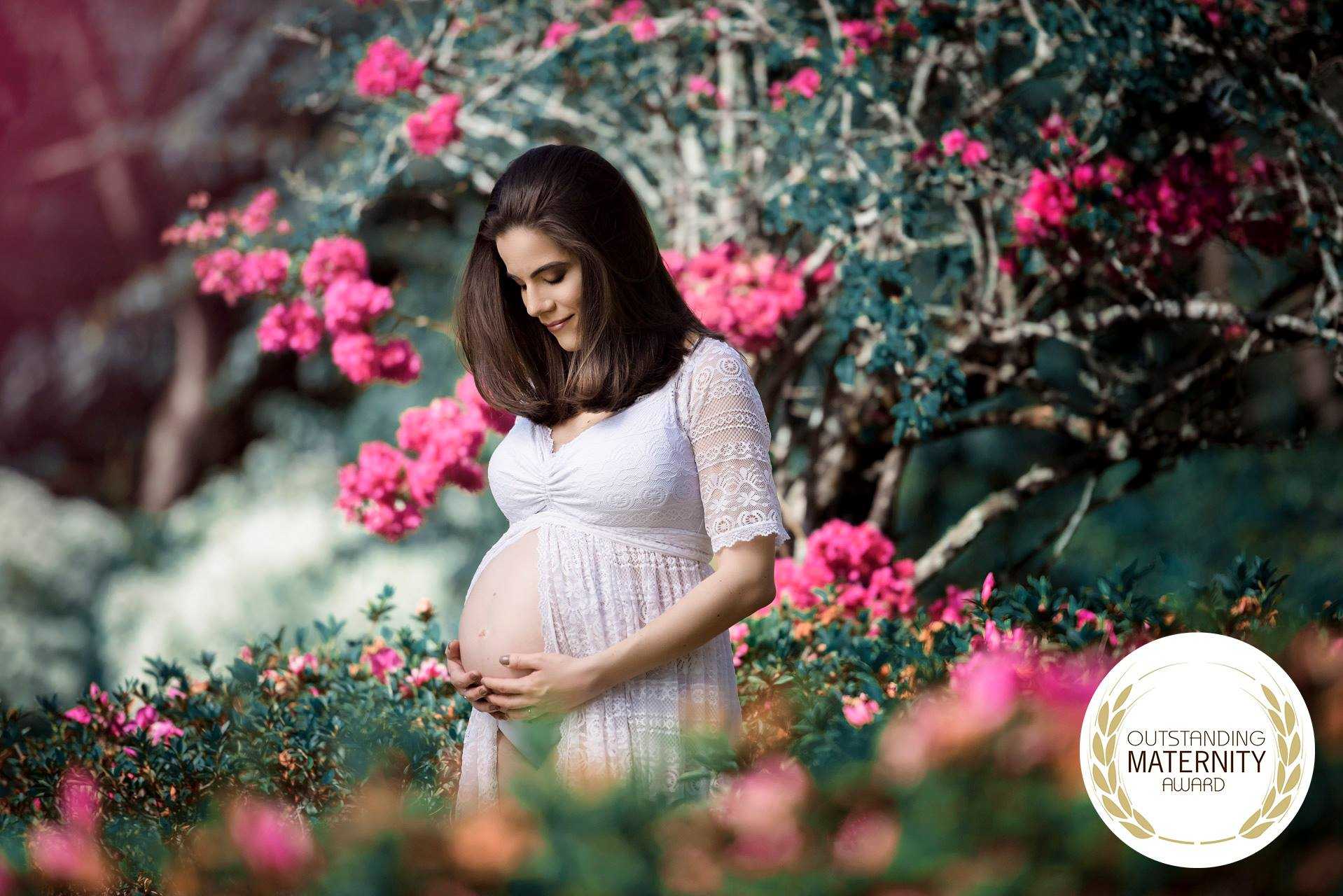 Foto de uma mulher grávida em um jardim florido, rodeada por flores rosa e verdejantes, simbolizando maternidade, vida nova e esperança